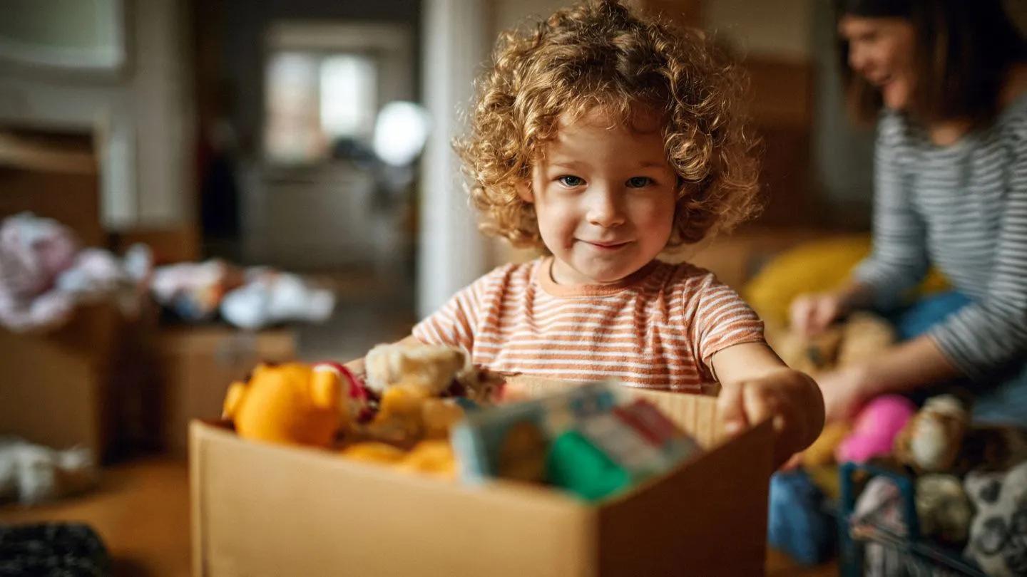 Smiling child with curly hair playing with toys in a cardboard box, indoors with an adult in the background.